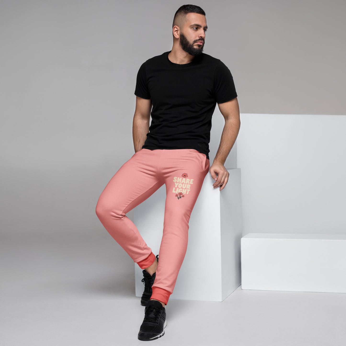 A man sitting on a white pedestal wearing peach-colored joggers with a graphic on the left leg and a black T-shirt.